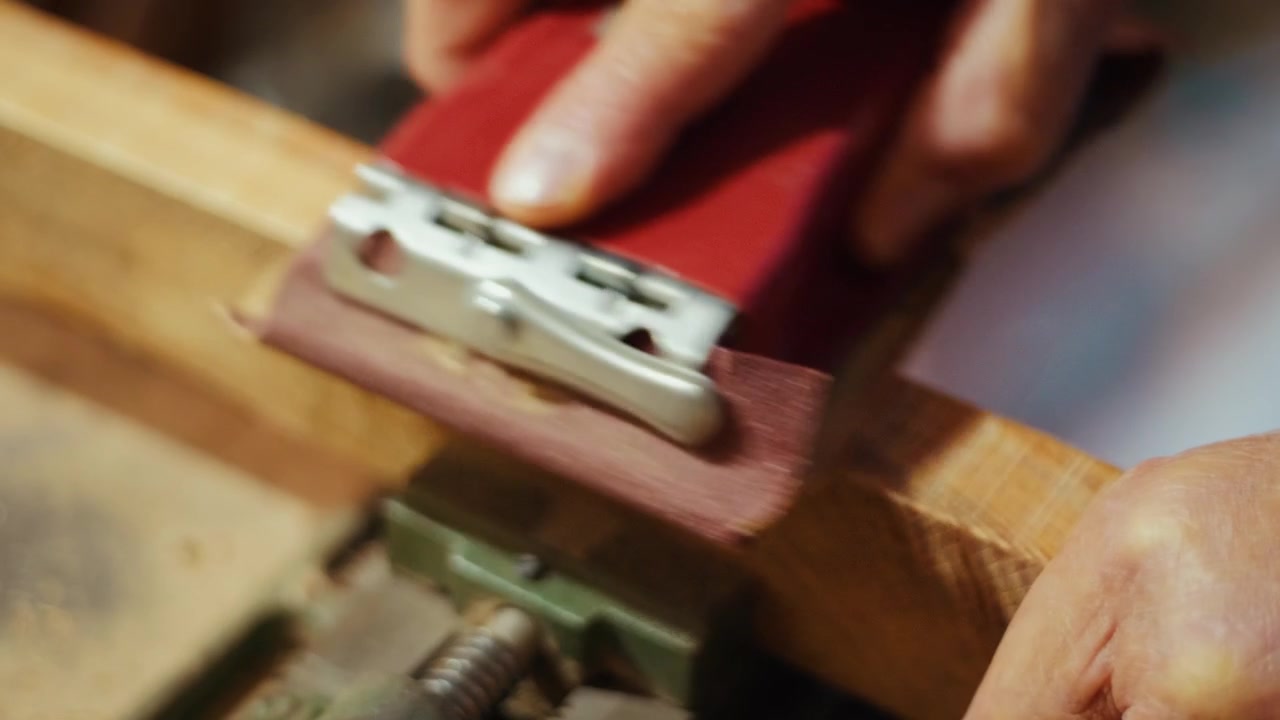 Close-up de um artesão usando um bloco de lixa vermelho para alisar uma tábua de madeira manualmente em uma oficina. Imagens autênticas de marcenaria DIY para projetos de artesanato, carpintaria e criação