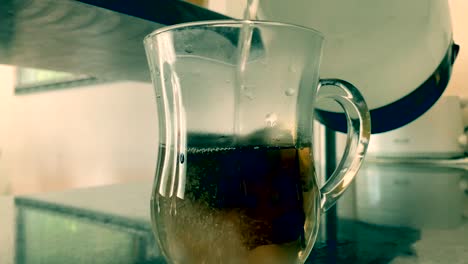 pouring boiling water from a glass pitcher into a cup with teabags to prepare the tea beverage, close up still shot