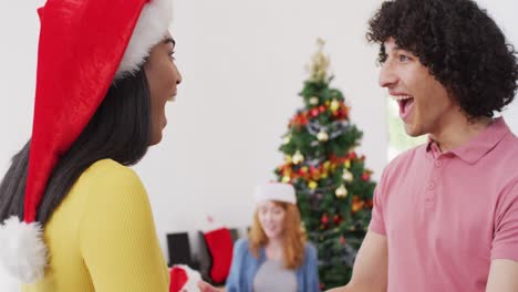 Happy-biracial-man-giving-present-and-hugging-non-binary-transgender-friend-in-santa-hat-at-home