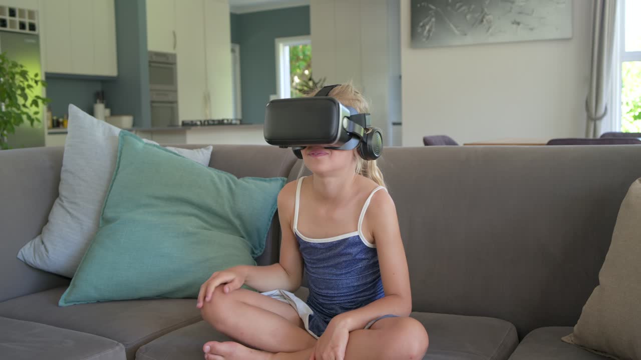 School-age girl in blue top responding to VR headset prompts, reaching, turning, reacting on sofa