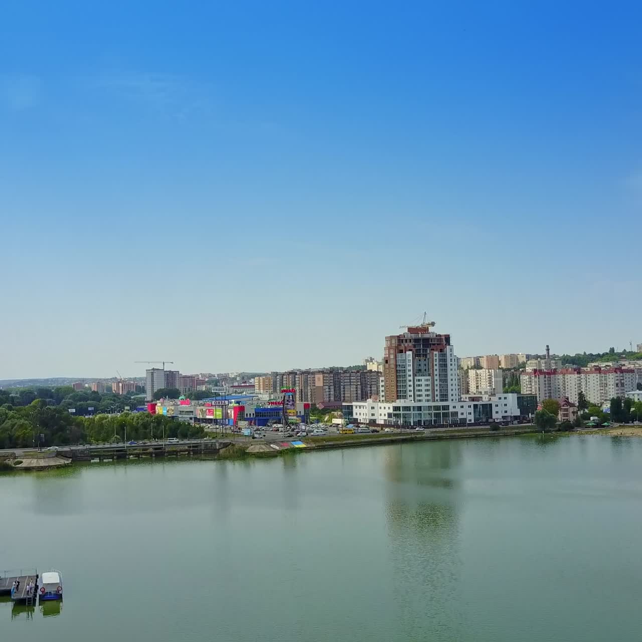 Flying above the river floating through the modern city. Lively traffic by the roads at waterfront among the multi-storied buildings. Top view