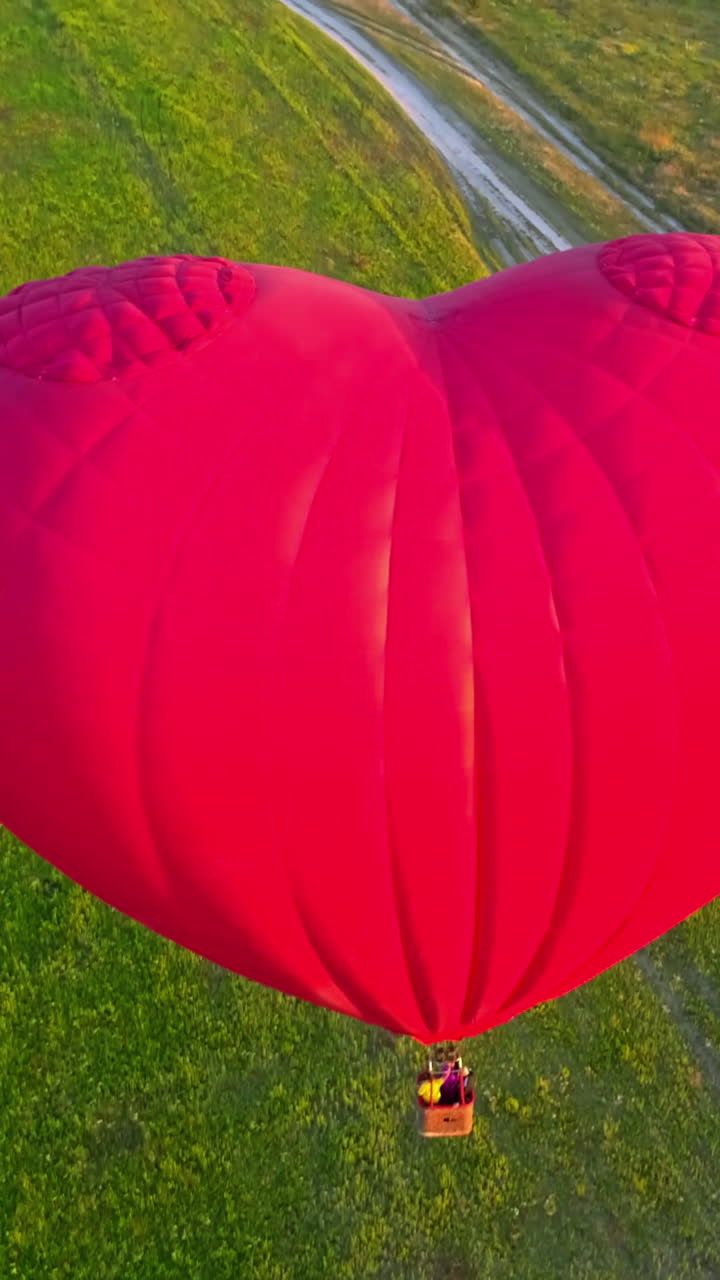 Large aerostat in the air. Beautiful red hot air balloon in the heart shape flying in the countryside. People travel in the basket of the airship in summer. Close-up. Vertical video