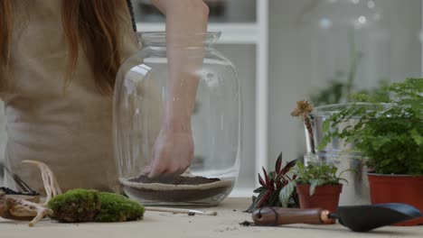 A-young-woman-carefully-puts-layers-of-soil-and-sand-into-a-glass-jar-and-looks-into-it---close-up