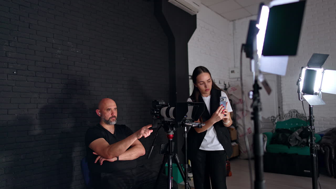 Caucasian photographer sits at camera on tripod. Brunette woman using phone for taking video. Studio footage.