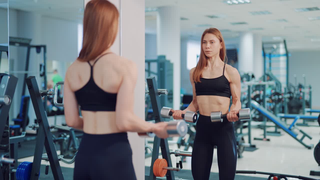 A beautiful woman in black leggings and top is doing an exercise for biceps using dumbbells opposite mirror in the gym. Blurred Background