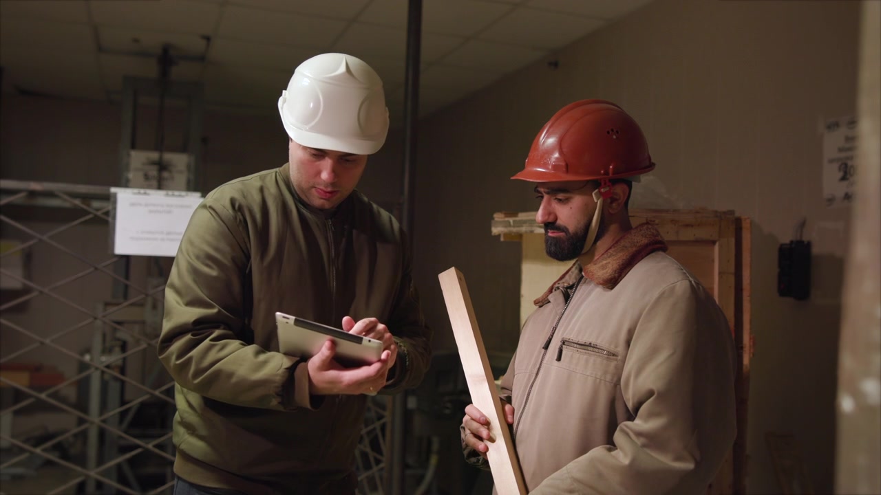 Warehouse Workers Inspect Plank And Smartphone, Lowlit Storage Room With Stacked Boxes And Scaffolding, Hard. Media