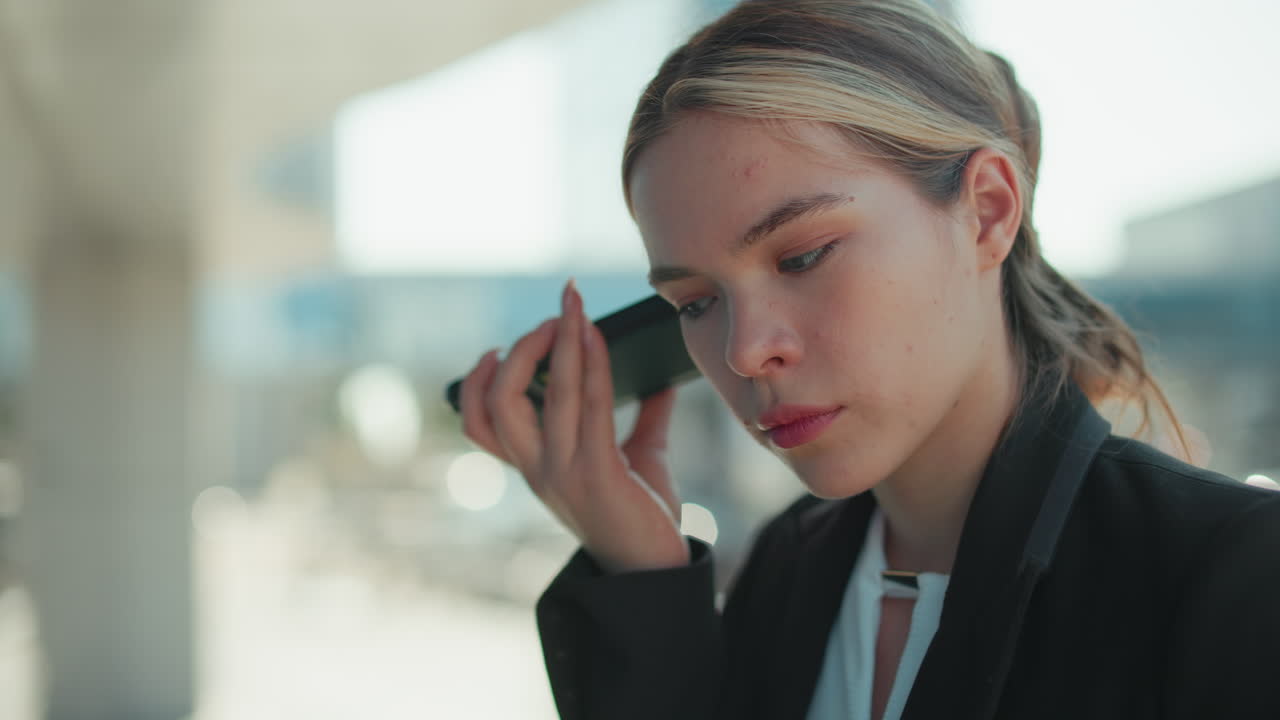 Close up of female lawyer attentively listening to voice note from client while standing outside modern glass building reflecting surroundings