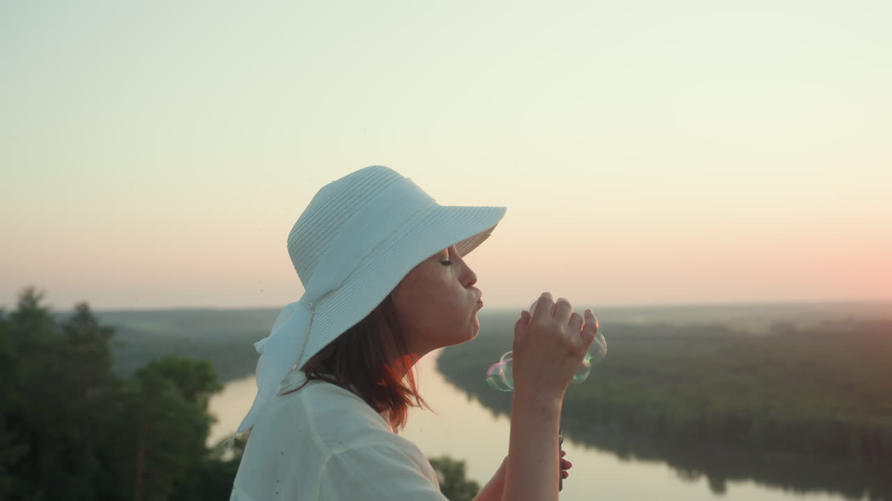 Close up side view of lady in white sun hat gently blowing soap bubble near river bend at golden hour with warm sunlight casting soft glow over forested landscape