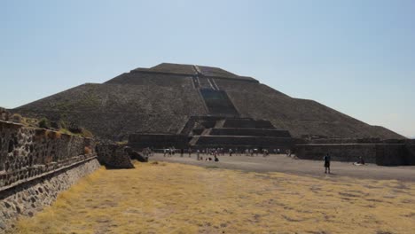 a panning wide and slow motion shot of the pyramid of the sun at the teotihuacan archaeological site in mexico, with some tourists walking around and the blue sky on a clear and sunny day