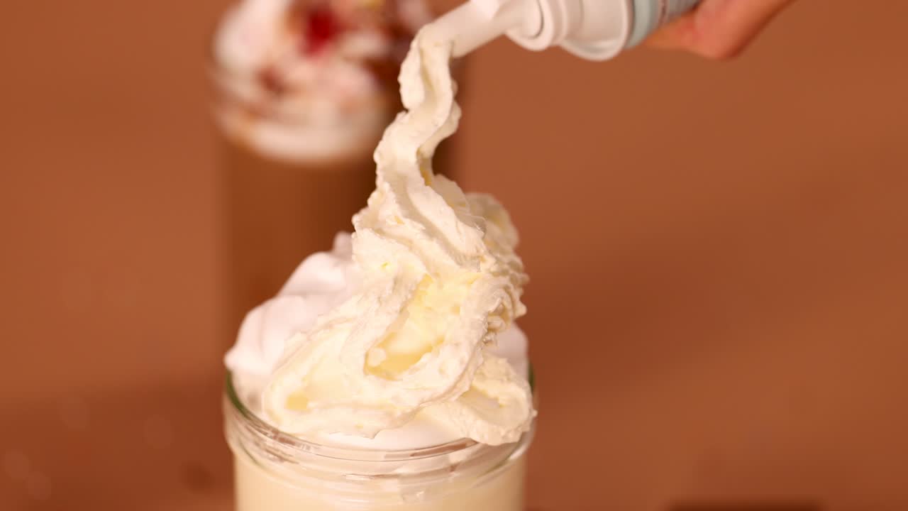 Hand adds whipped cream topping to creamy vanilla milkshake in glass jar, warm brown background