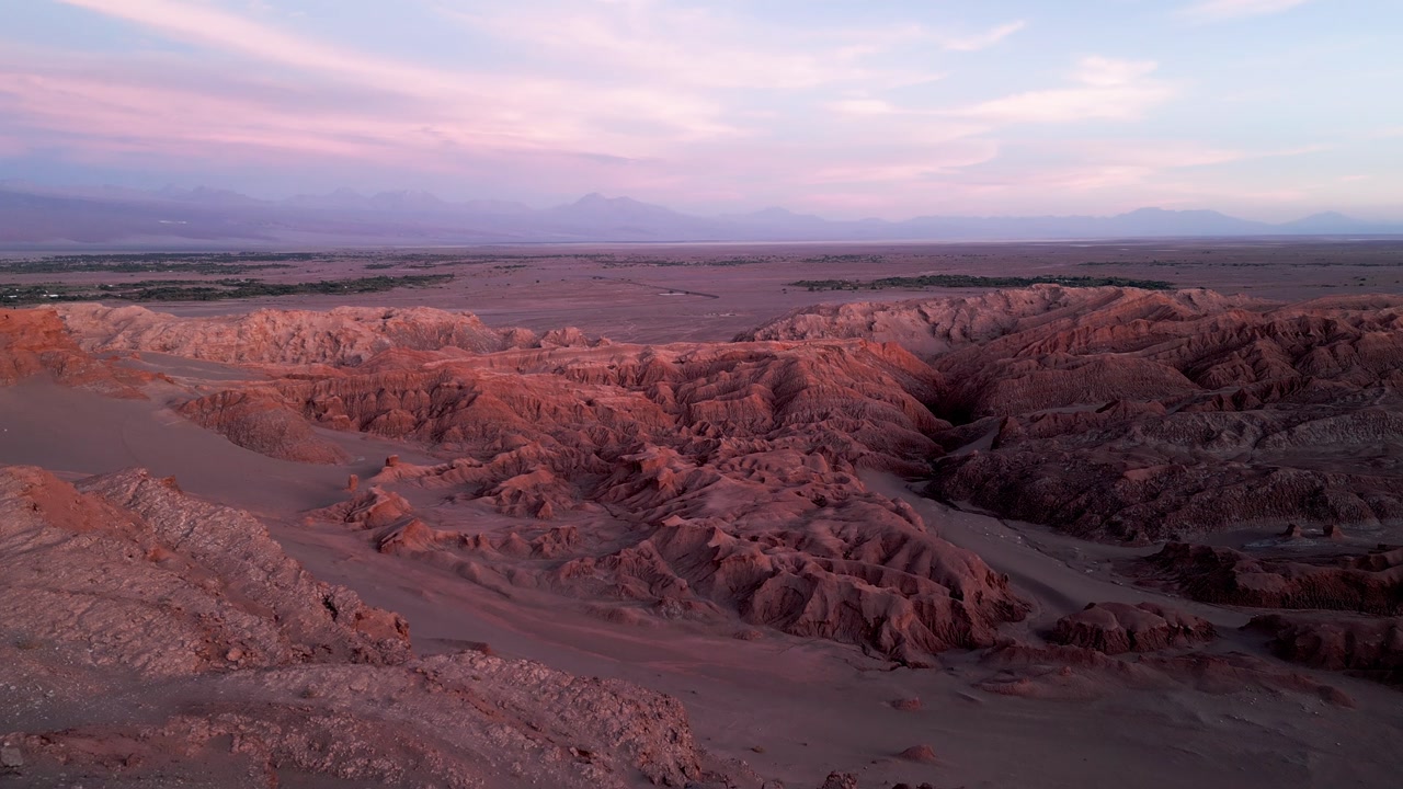 Backward drone flight over dramatic eroded desert canyon badlands at sunset revealing the vast Atacama plateau oasis and distant Andean peaks under a pink and purple sky. Valle de la Luna, San Pedro d