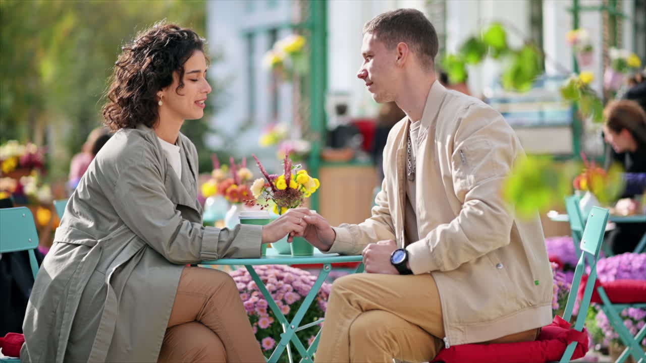 A romantic couple outdoors near a cafe. Holding hands, smiling, talking and looking at each other, flowers on the table. Autumn atmosphere. Slow motion