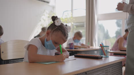 student in protective mask studying at school during corona virus pandemic. a masked teacher explains a new lesson topic to masked students. children write with pens in the classroom