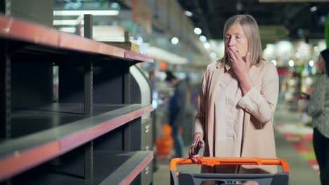 senior woman shopper in supermarket, grocery store near empty shelves counters. elder mature female panic buying and stockpiling. groceries supplies with sold out products.