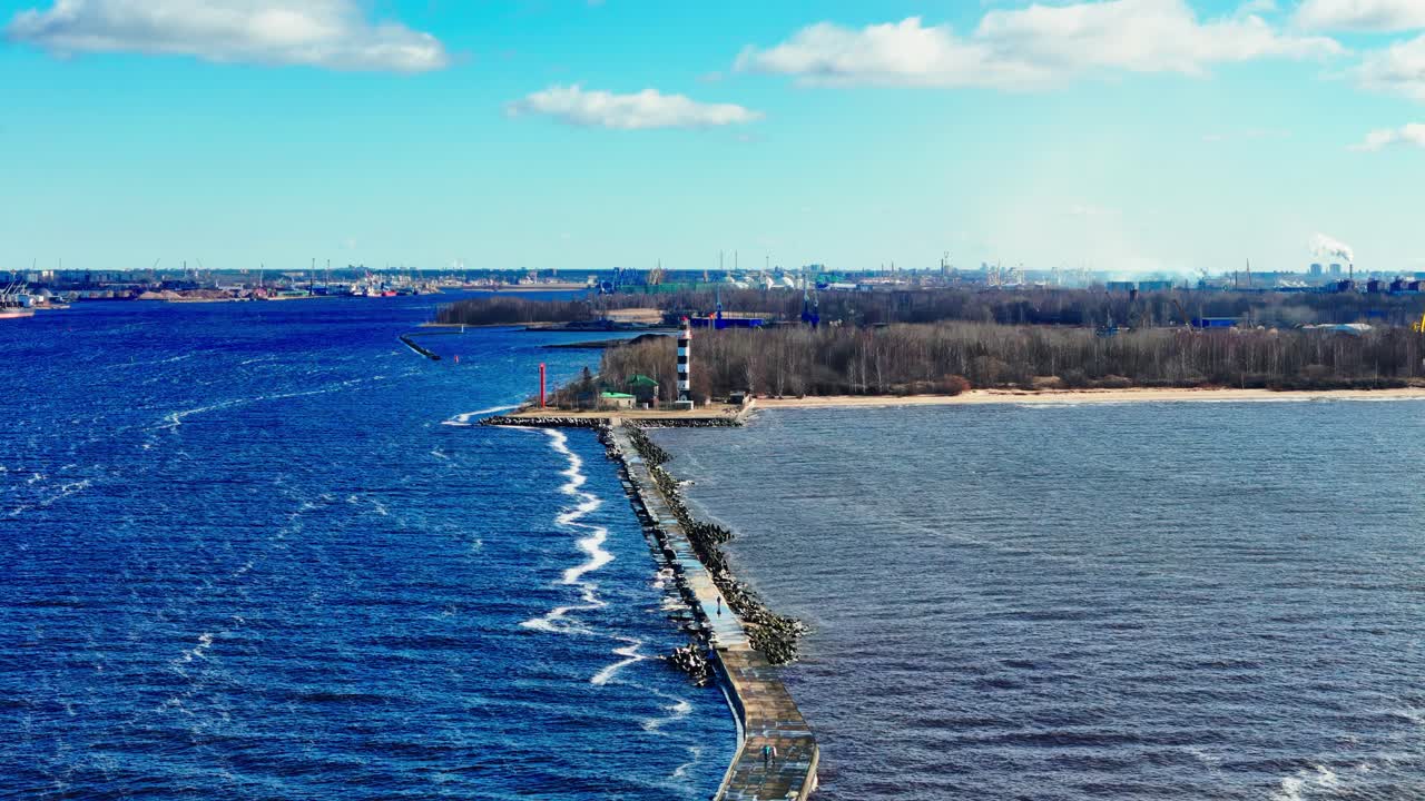 A long stone jetty with a tall black and white lighthouse divides vivid blue Baltic Sea from muted estuary waters, bordered by sandy shore, forest, and distant industrial skyline.