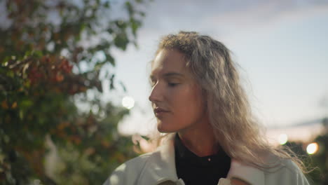 white lady walking along green path touching plant leaves gently while admiring nature with soft sunlight reflecting on face during peaceful evening stroll under colorful sky with blurred background