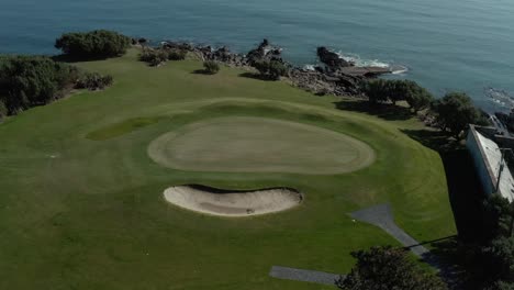 Vista-Aérea-De-Pájaro-Del-Campo-De-Golf-Junto-Al-Océano-En-Calma-Durante-El-Día,-Sin-Gente