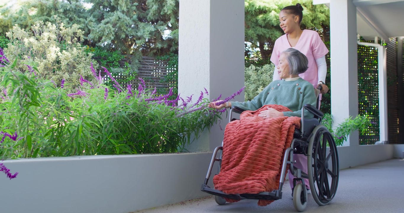 Assistente donna che spinge un anziano asiatico su una sedia a rotelle, passando accanto a fioriere in fiore, l'anziano che allunga la mano, spazio per il testo.