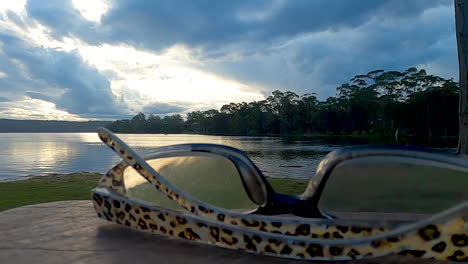 las gafas de lectura se colocan en una mesa de picnic con vistas a un lago al atardecer en una noche nublada