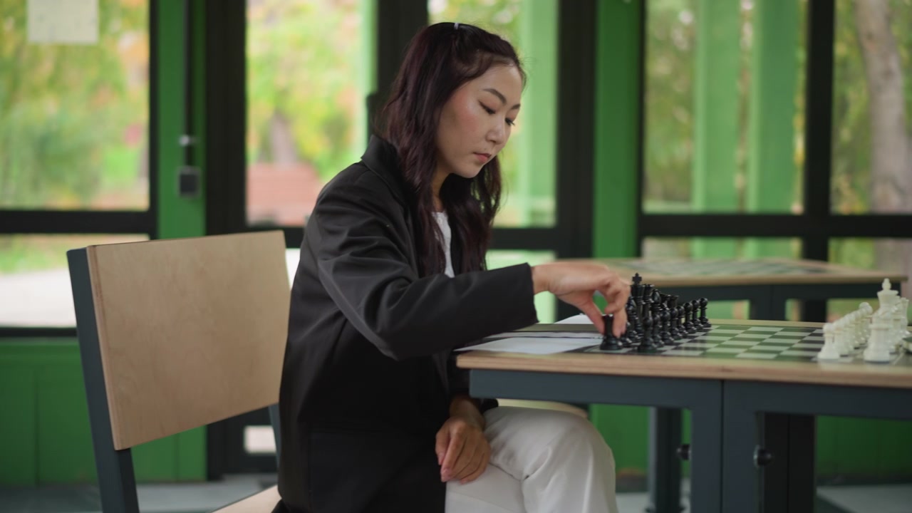 Close-up seorang wanita muda bermain catur di meja dengan buku catatan, tenggelam dalam pikiran tentang langkah selanjutnya. Wanita mengenakan blazer hitam dan celana putih saat duduk di kursi kayu dekat jendela besar.