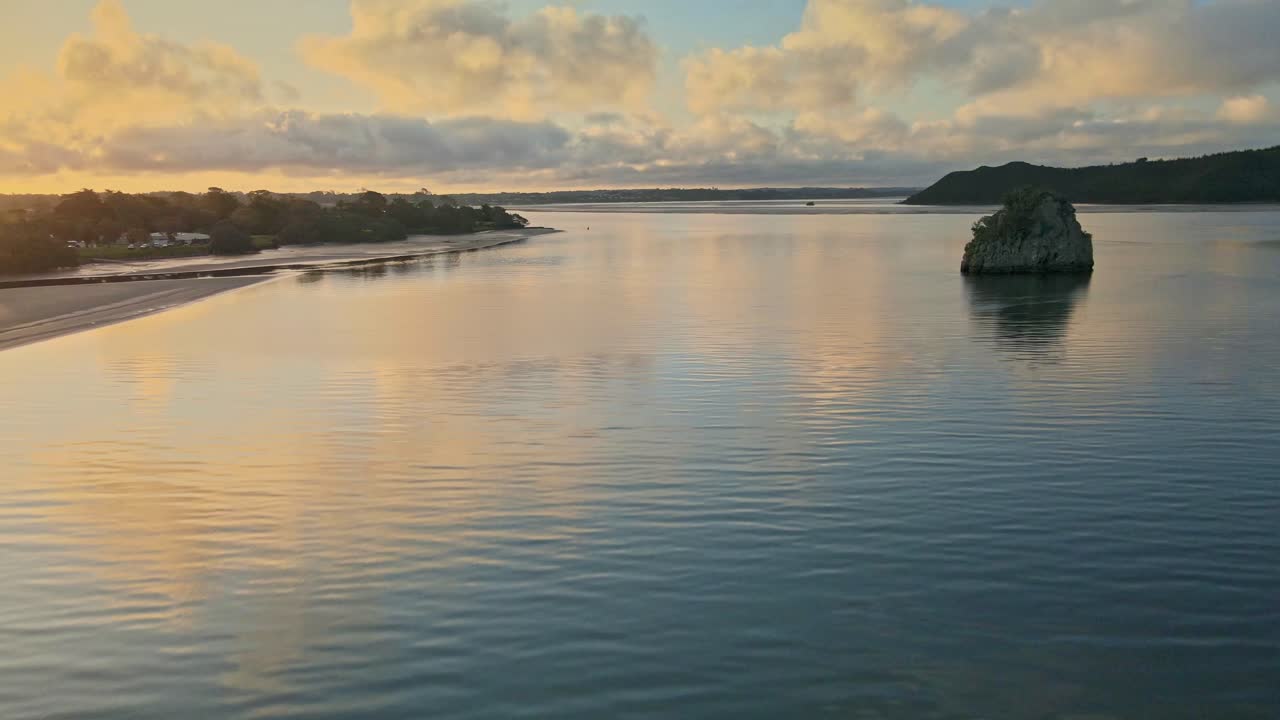 Gentle drone glides over calm Pukenui waters, where the ocean reflects golden sunset light, clouds glow with color, creating a serene, tranquil and peaceful coastal atmosphere in New Zealand