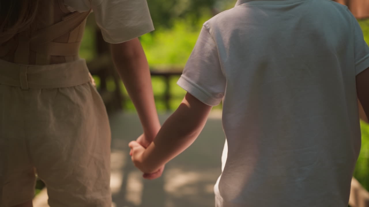 Little girl holds toddler boy hand walking together along overpass in forest closeup. Children enjoy summer and friendship at countryside nature