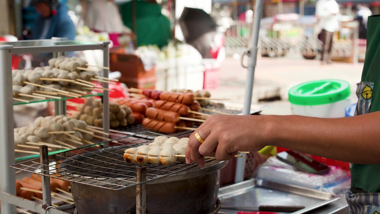 Primo piano di un venditore che griglia spiedini assortiti di polpette e salsicce al Mercato dei Fiori di Bangkok, Thailandia