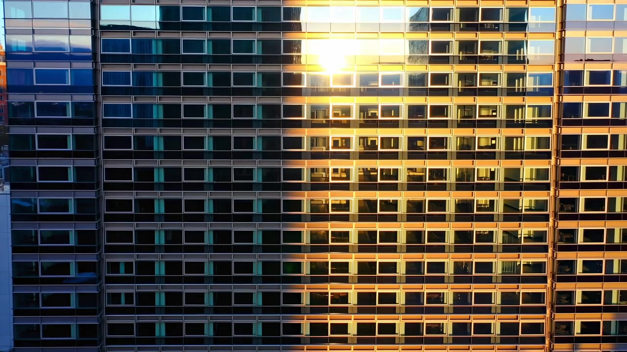 A luz do sol se move pela fachada de um edifício de escritórios alto, mudando de uma sombra azul-petróleo fria para um reflexo dourado quente e criando faixas verticais crescentes de reflexos brilhantes nos painéis de vidro