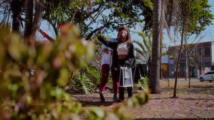 Two women taking a selfie outdoors in a park-like setting after shopping