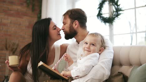 El-Joven-Padre-Y-Su-Hijo-Leen-Un-Libro,-Mientras-La-Madre-Bebe-Té-Y-Besa-A-Su-Marido.-Familia-Leyendo-Cuento-De-Navidad