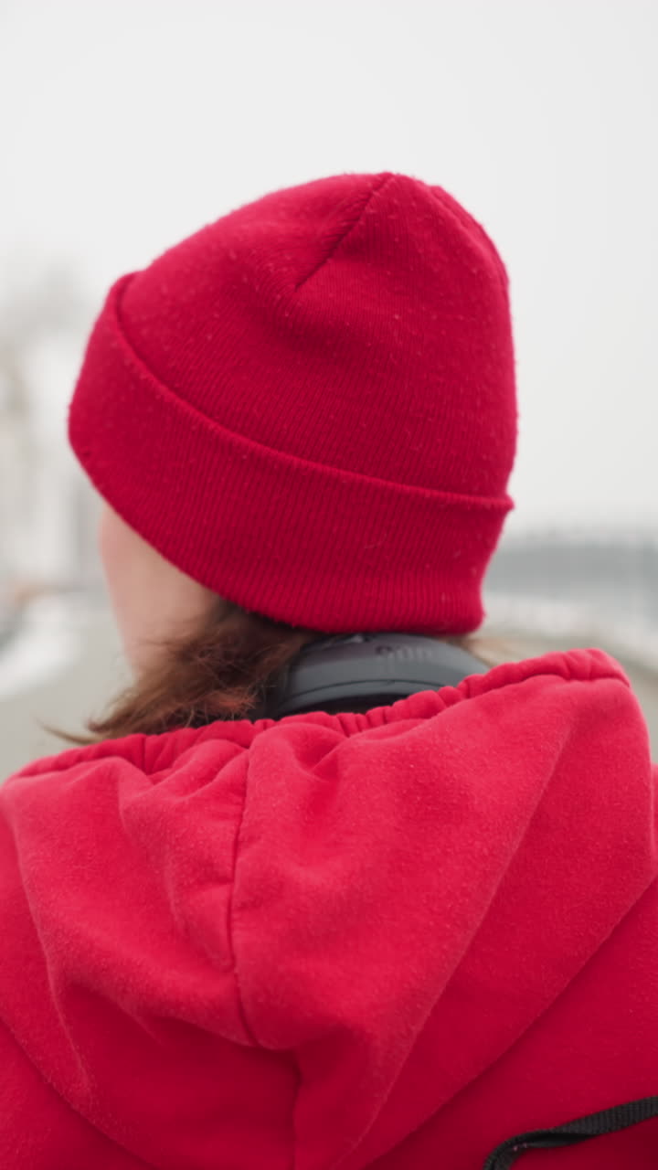 은 베니와 재을 입고 목에 헤드폰을 달고 은 나무와 램프 기둥을 배경으로 눈 인 겨울 공원 설정에서 철 레일링 근처의 인 경로를 고 있는 여성의 뒷면