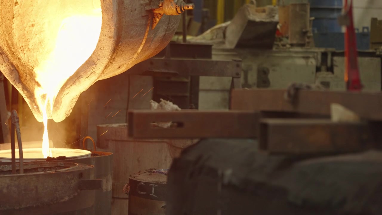 White hot molten steel flows from a tilting crucible into a round casting mold on a busy foundry floor.