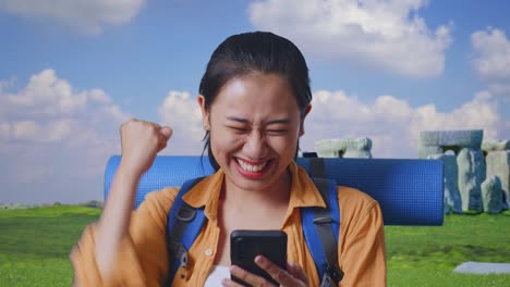 close up of asian female hiker with mountaineering backpack looking at the smartphone and screaming goal celebrating the success while traveling in stonehenge