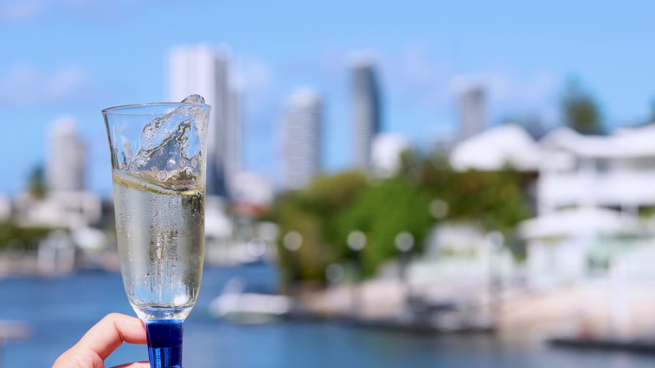 A hand swirls a champagne glass, causing bubbles and liquid motion, with a sunlit waterfront cityscape and modern buildings softly blurred in the background