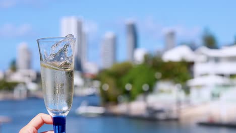 a hand swirls a champagne glass, causing bubbles and liquid motion, with a sunlit waterfront cityscape and modern buildings softly blurred in the background