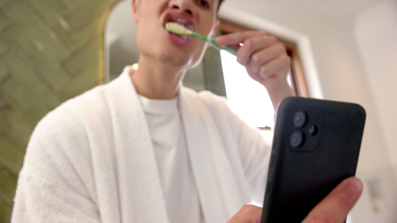 Homem birracial escovando os dentes e usando o smartphone no banheiro pela manhã, em câmera lenta.