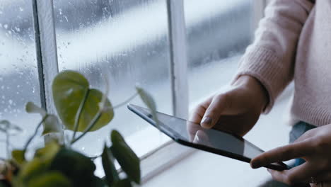 Cerrar-Las-Manos-De-Una-Mujer-Usando-Una-Tableta-Digital-Navegando-Mensajes-En-Línea-Leyendo-Las-Redes-Sociales-Disfrutando-De-Un-Dispositivo-Móvil-Con-Pantalla-Táctil-Parado-Junto-A-La-Ventana-Relajándose-En-Casa-En-Un-Día-Frío-Y-Lluvioso