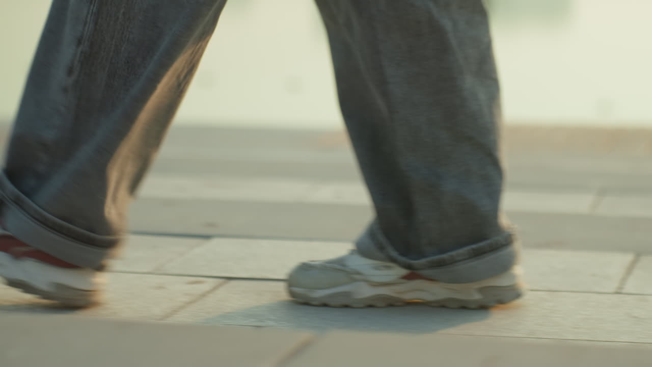 Primer plano de pies caminando sobre pavimento de un viajero urbano, zapatillas gastadas y vaqueros holgados, paso lento y deliberado por una plaza embaldosada, luz dorada cálida, sombras suaves, piedra texturizada, paso rítmico, cinematográfico