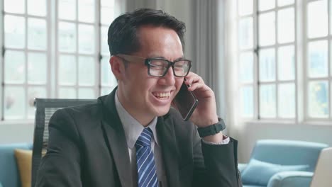 Asian Businessman With Glasses Wearing Business Suit Answering The Phone Call While Using The Computer For Working At Home.