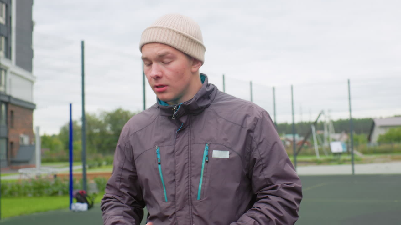 white man shoots basketball on court, wearing knit beanie and insulated jacket, overcast sky, fenced outdoor court in suburban setting, focused expression, midshot showing ball motion toward hoop