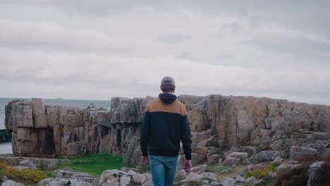 Young-Man-In-Jacket,-Cap-and-Jeans-Walking-Towards-Vårhallarna-Cliffs---Medium-Wide-Shot-Tracking-Forward