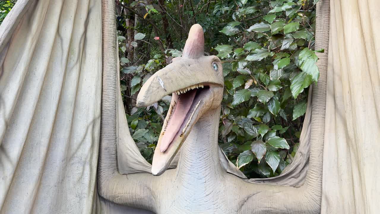 Animatronic pterosaur turns head, opens mouth in bright, leafy outdoor setting, mid-range shot