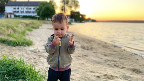 beautiful caucasian baby boy stands on the beach looking at his hands dirty with sand. kid claps his hands to clean them.