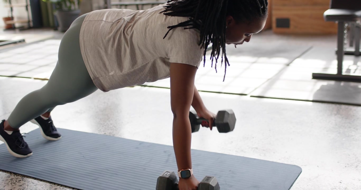 Mid-adult woman of African descent grabbing hex dumbbell in gym planking alternating rows for core