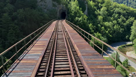 an old train railway crosses a weathered wooden bridge, emerging from a dark tunnel and winding through a dense mountain forest, wide daytime tracking shot