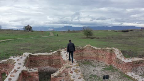 aerial view of a man walking through ancient city ruins in a large green field on a cloudy day