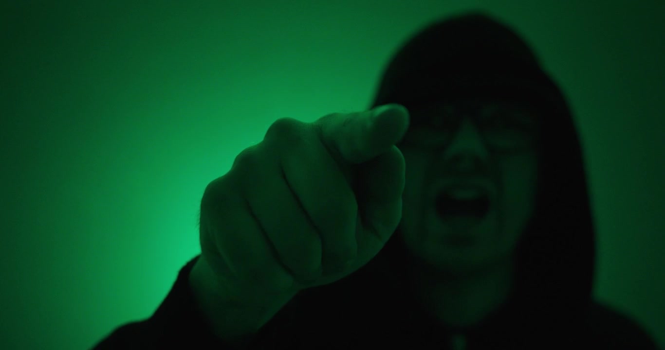 Uomo incappucciato che punta direttamente verso la telecamera sotto un'illuminazione verde drammatica. Concetto di accusa, avvertimento, mistero, suspense, confronto e narrazione cinematografica
