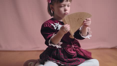 cute little girl in an vintage burgundy dress is playing with a wooden fan