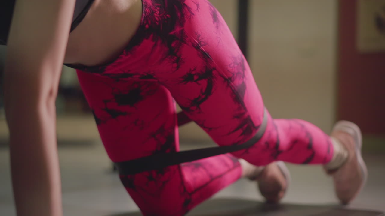 donna in leggings rosa che esegue kickback di resistenza su tappetino con fascia ad anello, primo piano dal basso che mostra l'attivazione dei glutei, ritmo controllato, illuminazione da studio, espressione determinata, focalizzata sulla forza