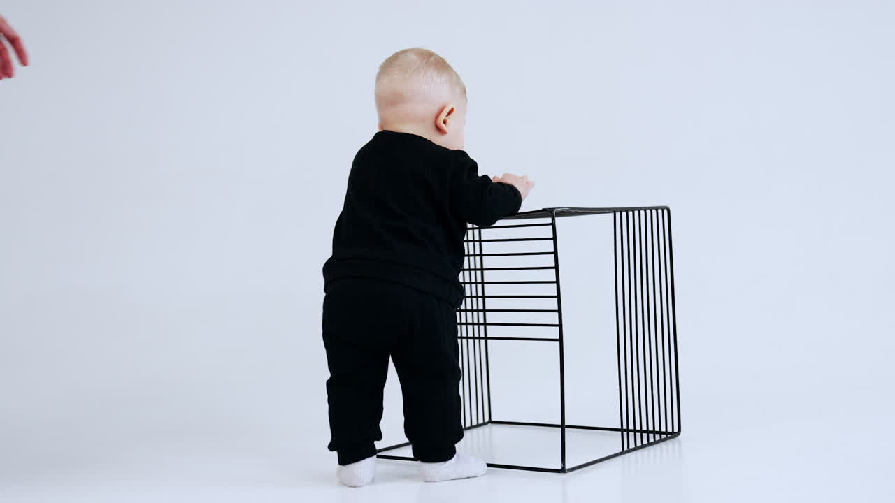 Lovely baby boy in black suit stands at the metal grate cube. Man comes up to child trying to turn him to camera. White backdrop.